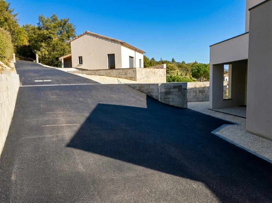 Allée privée en enrobé bitumineux noir, fraîchement posée, desservant deux maisons individuelles contemporaines en enduit clair