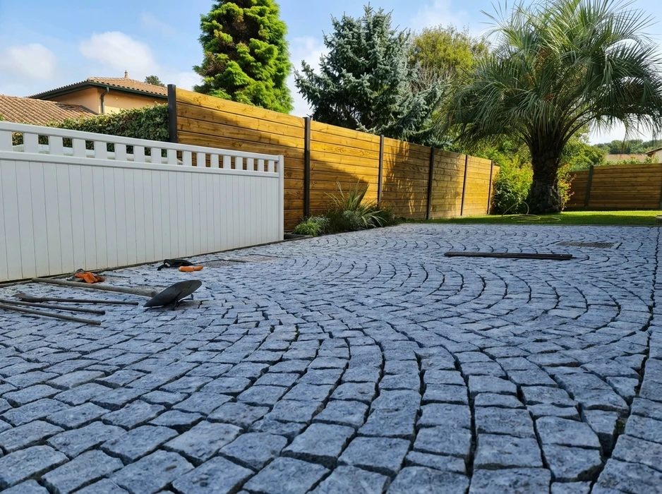 Ouvrier en équipement de sécurité orange et casque blanc installant du pavage extérieur, agenouillé sur dalle béton, utilisant outil de coupe sur pavés gris